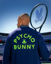 On a tennis court, a man holds his racket over his shoulder, showing the back of his Psycho Bunny TENNIS BALL CREW MENS JACKET ENGLISH, detailed with neon yellow "PSYCHO BUNNY" text and an embroidered Bunny logo with crossed rackets.
