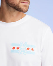 A man wears the Psycho Bunny MENS CHICAGO CITY TEE - B6U551W1PC, a white Pima cotton shirt featuring a blue and red rabbit skull and crossbones graphic with red stars on the chest, set against a light blue background for a vibrant look.