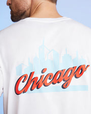 A person in a white MENS CHICAGO CITY TEE by Psycho Bunny displays a red "Chicago" cursive graphic over a light blue city skyline. They are partially turned away, showing just part of their face.