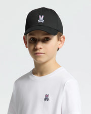 A young person wears a Psycho Bunny black KIDS CLASSIC BASEBALL CAP (B0A816A1HT) with an embroidered bunny and crossbones, complemented by a white T-shirt featuring the same bold logo. The plain white background accentuates their edgy style as they look at the camera.