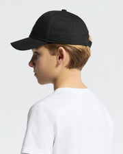 A youth wearing the Psycho Bunny KIDS CLASSIC BASEBALL CAP in black, product code B0A816A1HT, with an adjustable fit and an embroidered Bunny logo on the left side, paired with a classic white T-shirt against a simple light-colored background.