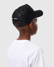 A child is seen from behind at an angle, wearing a Psycho Bunny KIDS BUNNY PATCH TRUCKER CAP (B0A965C200) with black breathable mesh paneling and a white t-shirt against a plain light gray background.