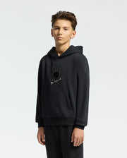 A young person stands against a plain background, wearing a black Psycho Bunny KIDS NEW CASTLE EMBROIDERED FRENCH TERRY HOODIE (B0H517F200) featuring an embroidered bunny and crossed bones logo. Their short brown hair frames their face as they gaze at the camera with a neutral expression.