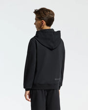 A person with short hair is wearing a black French terry KIDS NEW CASTLE EMBROIDERED HOODIE by Psycho Bunny, along with black pants, standing and facing away. The background is plain light gray.