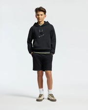 A teen sports the Psycho Bunny Kids New Castle Embroidered French Terry Hoodie - B0H517F200, teamed with black shorts, white socks, and gray sneakers. Their hands are tucked in pockets against a plain white background.
