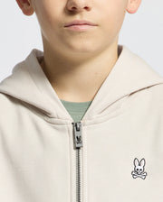 A person models the KIDS LEVY ESSENTIAL ZIP FLEECE HOODIE (Beige, B0H720D200) by Psycho Bunny, featuring a small embroidered bunny with crossbones. The light, plain background adds mystery as only the mouth and part of the nose are visible.