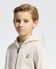 A young person with short, light brown hair is wearing a light-colored Kids Levy Essential Zip Fleece Hoodie by Psycho Bunny, featuring a small rabbit emblem on the chest, standing against a plain background.
