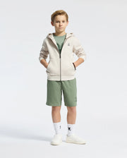 A young boy stands with his hands in the pockets of a Psycho Bunny beige fleece hoodie. He's wearing a green t-shirt, green shorts, white socks, and white sneakers against a plain white background.