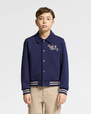 A young boy with short brown hair stands facing forward, wearing the Psycho Bunny KIDS DESERT COACH BOMBER JACKET (B0J428F200) in Evening Blue with striped cuffs and waistband, cursive chest text, and beige pants against a plain white background.