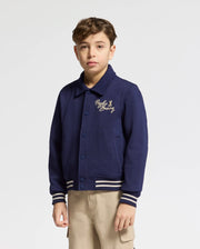A young boy with short brown hair wears the Psycho Bunny KIDS DESERT COACH BOMBER JACKET (B0J428F200) in Evening Blue, featuring striped cuffs and gold chest embroidery, paired with beige pants against a plain white background.