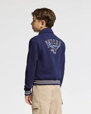 A young boy faces away in a Psycho Bunny KIDS DESERT COACH BOMBER JACKET (B0J428F200) in Evening Blue, featuring "PSYCHO Bunny" and a bunny logo on the back, paired with beige cargo pants against a plain white background.