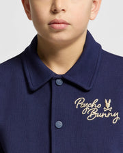 A child wears the Psycho Bunny KIDS DESERT COACH BOMBER JACKET (B0J428F200) in Evening Blue, featuring "Psycho Bunny" and a beige bunny logo embroidered on the left chest. The double-knit jacquard fabric adds subtle texture; only part of the face shows.