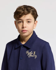 A young boy with short brown hair wears the Psycho Bunny KIDS DESERT COACH BOMBER JACKET (B0J428F200) as he stands against a plain light background.