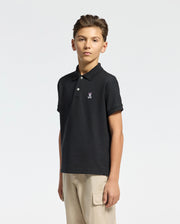 A young person stands against a light background wearing the KIDS CLASSIC PIQUE POLO SHIRT (B0K001CRPC) by Psycho Bunny in black, crafted from soft Pima cotton with the classic logo. They pair it with beige cargo pants, have short hair, and maintain a neutral expression.