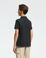A young person with short hair faces away, wearing the KIDS CLASSIC PIQUE POLO SHIRT in black and beige pants. Featuring the subtle Psycho Bunny logo, this soft Pima cotton piece is set against a plain, light backdrop.