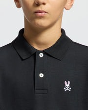 The image shows someone in a Psycho Bunny KIDS CLASSIC PIQUE POLO SHIRT - B0K001CRPC, made of soft Pima cotton, with two buttons fastened. The black shirt features the classic logo: a pink-eared bunny above white crossed bones. The background is plain and light, with part of the person's face visible.