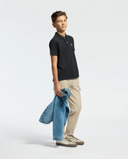 A teen boy wears a KIDS CLASSIC PIQUE POLO SHIRT - B0K001CRPC by Psycho Bunny, paired with beige pants. He holds a denim jacket in his left hand, sports light-colored sneakers, and gazes slightly to his right while standing on a plain background.