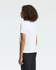A person with curly hair, seen from the side and facing away, wears a white Psycho Bunny Kids Classic Pique Polo Shirt (B0K001CRPC) paired with black pants against a plain background.
