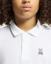 A person with curly hair is wearing a Psycho Bunny KIDS CLASSIC PIQUE POLO SHIRT in soft Pima cotton, adorned with a small pink bunny logo and crossbones on the left side. The image shows their upper torso and face, excluding the eyes.