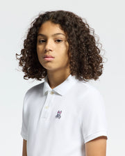 A person with curly hair dons a Psycho Bunny KIDS CLASSIC PIQUE POLO SHIRT - B0K001CRPC, made from soft Pima cotton and featuring an embroidered logo on the chest. The light color background enhances its timeless style.
