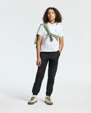 Curly-haired, wearing a Psycho Bunny Kids Classic Pique Polo Shirt in white with black pants and beige sneakers. A light green sweater drapes over their shoulders against a plain white background, hands casually in pockets.