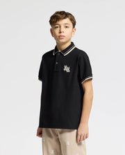 A young boy wears the Psycho Bunny KIDS ELLIOT PIQUE POLO in black Pima cotton, featuring an embroidered Bunny logo on the chest. His khaki shorts match with a similar design. The shirt's collar and sleeves sport white stripes on a white background.