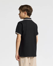 A boy with brown hair, seen from the back, wears the Psycho Bunny KIDS ELLIOT PIQUE POLO in black Pima cotton with white trim and khaki pants. The shirt features an embroidered bunny on the chest against a plain white background.