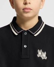 A child wears a Psycho Bunny KIDS ELLIOT PIQUE POLO in black, featuring soft Pima cotton with white trim on the collar and button placket. The standout design is an embroidered logo: two bunny heads crossed over bones on the chest, set against a plain light background.