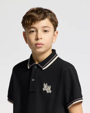 A young boy with short hair wears the Psycho Bunny KIDS ELLIOT PIQUE POLO (B0K505F200) in black, crafted from soft Pima cotton. It features an embroidered Bunny design on the chest and white trim on the collar and sleeves, contrasting against a light background.