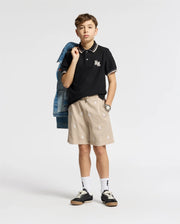 A young boy confidently wears a black KIDS ELLIOT PIQUE POLO - B0K505F200 by Psycho Bunny, beige patterned shorts, and black sneakers. He casually slings a denim jacket over his shoulder while sporting a stylish watch against the plain white background.
