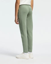 A person in Psycho Bunny's sage green KIDS DESERT EMBROIDERED MODAL SWEATPANT and a white T-shirt paired with white sneakers stands against a light background. The side angle view highlights the casual fit and soft stretch jersey material, enhanced by an embroidered wordmark graphic.