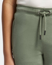 A person wears green KIDS DESERT EMBROIDERED MODAL SWEATPANT by Psycho Bunny, featuring a tucked-in white shirt. The focus is on the drawstring waistband and upper thigh area, showcasing the texture, fit, and embroidered wordmark detail.