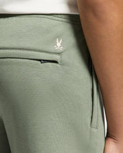A close-up displays the olive green KIDS DESERT EMBROIDERED MODAL SWEATPANT - B0P489F200 by Psycho Bunny, featuring a visible pocket and embroidered bunny logo above crossed bones near the waistband. Crafted from soft stretch jersey, they show a glimpse of a white shirt at the waist.