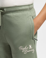 A person is wearing modal-blend sweatpants from Psycho Bunny, featuring the "Desert" embroidery near the pocket. These KIDS DESERT EMBROIDERED MODAL SWEATPANT (B0P489F200) are crafted from soft stretch jersey with an elastic waistband and drawstring, and they have a hand resting in the pocket.