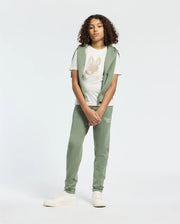 A teen is wearing a white T-shirt with an embroidered wordmark, a green sleeveless hoodie, and Psycho Bunny's KIDS DESERT EMBROIDERED MODAL SWEATPANT in green. The outfit features modal-blend sweatpants and soft stretch jersey material. Completing the look are white sneakers against a plain backdrop.