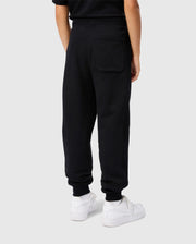 A person is standing against a light gray background, wearing Psycho Bunny's Kids Levy Essential Fleece Sweatpants (B0P721D200) in black, made from cozy fleece fabric with a large back pocket, paired with white sneakers. The upper body's not fully visible.