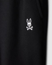 Close-up of the black fabric on Psycho Bunny's KIDS LEVY ESSENTIAL FLEECE SWEATPANT (B0P721D200), highlighting a white embroidered bunny skull and crossbones logo. The cozy fleece features a textured, ribbed look with a visible seam for added style.