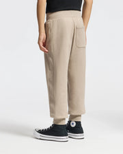 A person in Psycho Bunny's KIDS LEVY ESSENTIAL FLEECE SWEATPANT - B0P721D200 in beige, paired with black high-top sneakers with white soles and a circular patch logo, stands on a light background. The sweatpants feature an elastic waistband and a back pocket.
