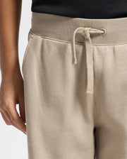 A person is wearing Psycho Bunny's KIDS LEVY ESSENTIAL FLEECE SWEATPANT - B0P721D200 in beige with a drawstring waist, paired with a black top. One hand rests by their side against a light background.