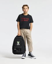 A young boy holds a black backpack with a cartoon skull and bunny logo, wearing a black Psycho Bunny T-shirt, beige KIDS LEVY ESSENTIAL FLEECE SWEATPANT - B0P721D200, and black high-top sneakers against a plain white background.