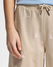A person in Psycho Bunny's KIDS ALL OVER WILLIS STRETCH TENCEL SHORTS, featuring tan fabric with white bunny and crossbones embroidery, exudes style. These regular-fit shorts have side pockets and are paired effortlessly with a light blue top.