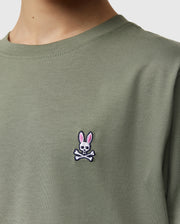 Close-up of a kid wearing the Psycho Bunny Kids Classic Crew Neck Tee in green. Perfect for back-to-school, this 100% Pima cotton tee features an embroidered bunny-headed skull and crossbones logo on the chest.