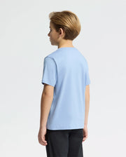 A young boy with short, light brown hair stands facing away from the camera, wearing a Psycho Bunny KIDS CLASSIC CREW NECK TEE (B0U014B200) in light blue with a small bunny logo, paired with dark pants against a plain, light gray background.