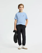 A boy wearing a light blue KIDS CLASSIC CREW NECK TEE by Psycho Bunny, made from soft Pima cotton with a subtle chest logo, holds a black bag with one hand. He is dressed in black pants and stands against a plain white background, looking forward.