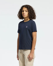 A young person with curly hair stands against a plain background, wearing a navy Kids Classic Crew Neck Tee by Psycho Bunny made from 100% Pima cotton and tan pants, exuding effortless style with their neutral expression and arms relaxed at their sides.