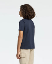 A person wears a Psycho Bunny Kids Classic Crew Neck Tee in navy blue with beige cargo pants, standing sideways with arms relaxed, looking left. Their curly hair enhances the casual vibe against a plain light background. This tee's Pima cotton offers both comfort and style effortlessly.