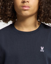 A person with curly hair wears a black Psycho Bunny KIDS CLASSIC CREW NECK TEE featuring a small pink bunny with crossbones logo near the chest. Their face is partially visible from the nose down.