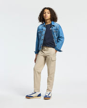 A person with curly hair wearing a classic denim jacket, a Psycho Bunny KIDS CLASSIC CREW NECK TEE in black, beige cargo pants, and blue and white sneakers stands confidently against a plain white background.