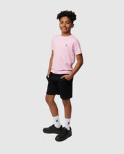 A young person stands confidently with hands in pockets, wearing a Psycho Bunny KIDS CLASSIC CREW NECK TEE - B0U014CRPC made from 100% Pima cotton. The pink tee is paired with black shorts, white socks featuring a small logo, and black shoes—ideal for stylish back-to-school outfits against the plain gray background.