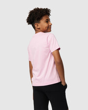 A young person with curly hair is wearing a light pink KIDS CLASSIC CREW NECK TEE from Psycho Bunny and black shorts. The 100% Pima cotton tee adds comfort to their back-to-school outfit as they stand smiling, looking over their shoulder against a plain, pale gray background.